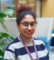 Dr Swarna Chetty a South Asian woman with dark curly hair tied up in a ponytail. She is wearing glasses and a striped top and is smiling at the camera.